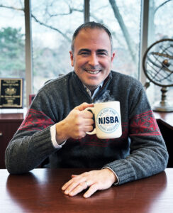 NJSBA CEO Tim Purnell, seated at desk, wearing a winter sweater, holding a white NJSBA coffee mug, smiling