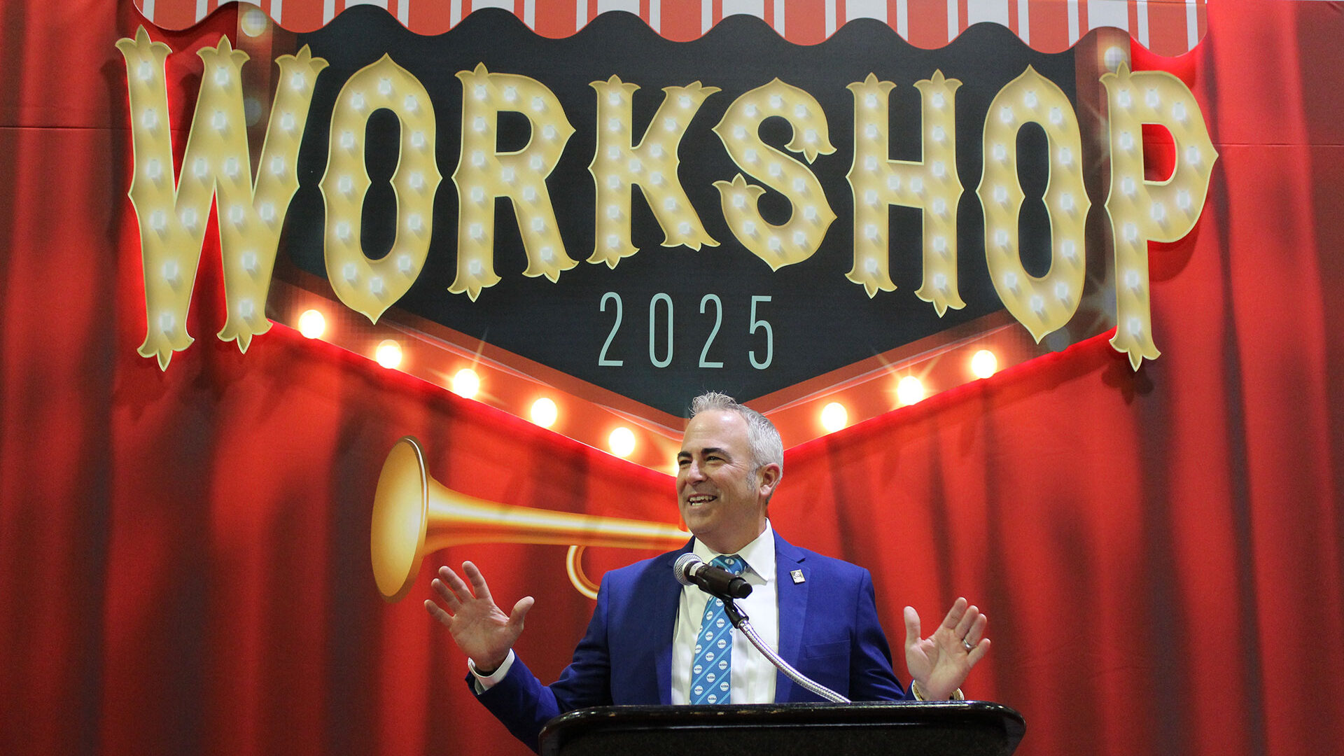 Dr. Timothy Purnell giving the opening ceremony speech in front of the workshop 2025 marquee light logo