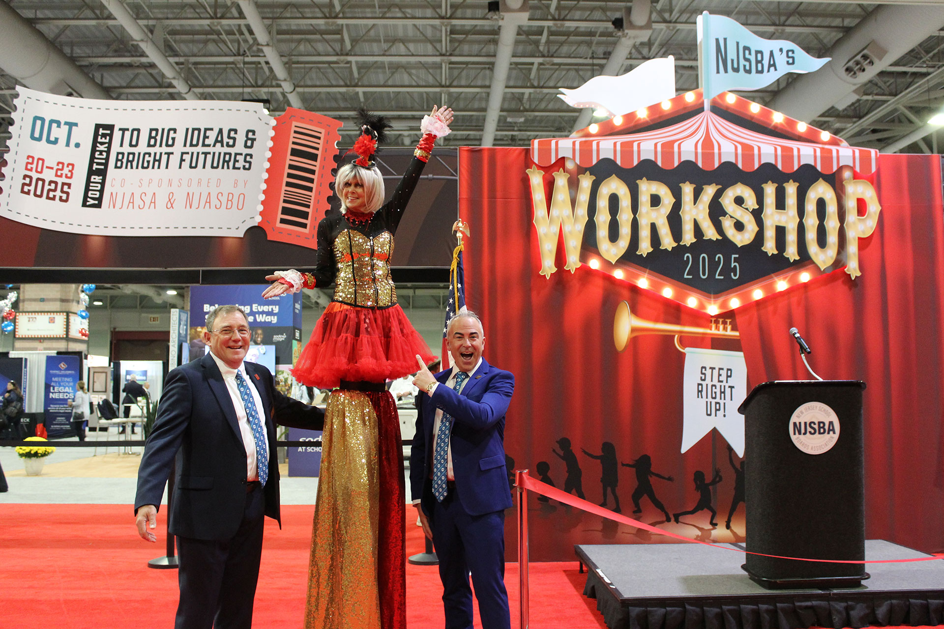 Dr. Timothy Purnel and Barry Walker posing with a women on stilts dressed in sequins in front of the workshop 2025 entrance unit