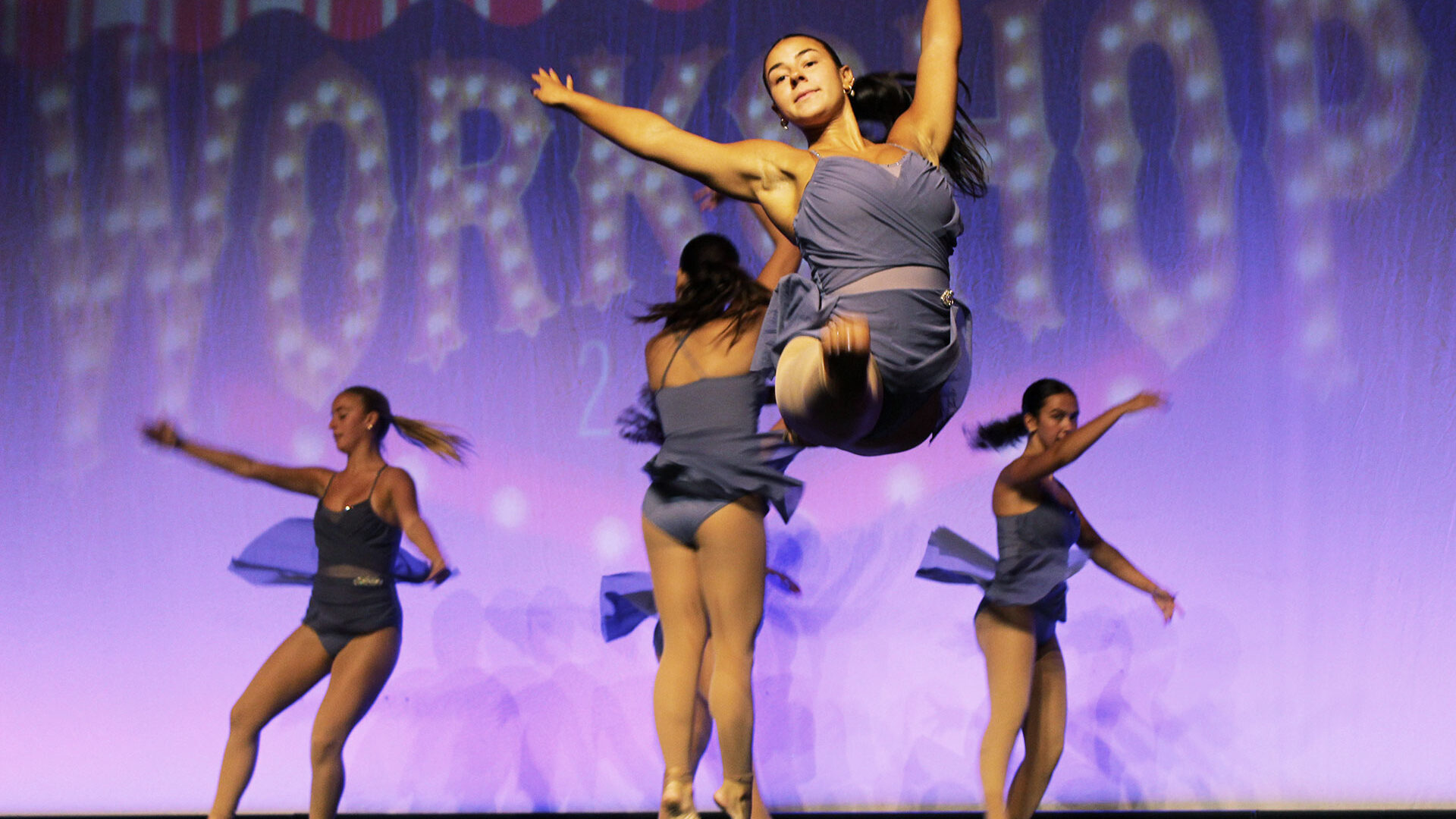 Student female dance team performing on a stage, one student leaping in the air doing a split