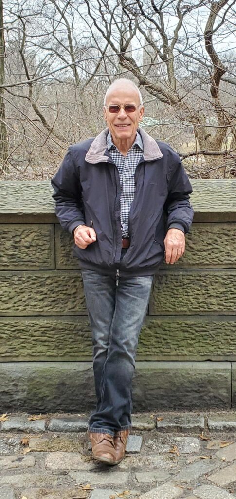2025 Unsung Superhero recipient Jay Aiello, dressed in blue collared shirt, navy blue jacket, sunglasses, and jeans, poses outside on a cloudy day.