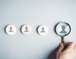 Four circles with headshot silloutte in the center are lined up. A hand holding a magnifying class is zoomed in on one