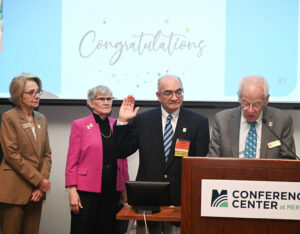 Michael Jacobs speaks while Charles Caravallo is sworn in, standing at a podium. Dr. Karen Cortellino and Irene LeFebvre stand beside. Congratulations is displayed on the screen behind.