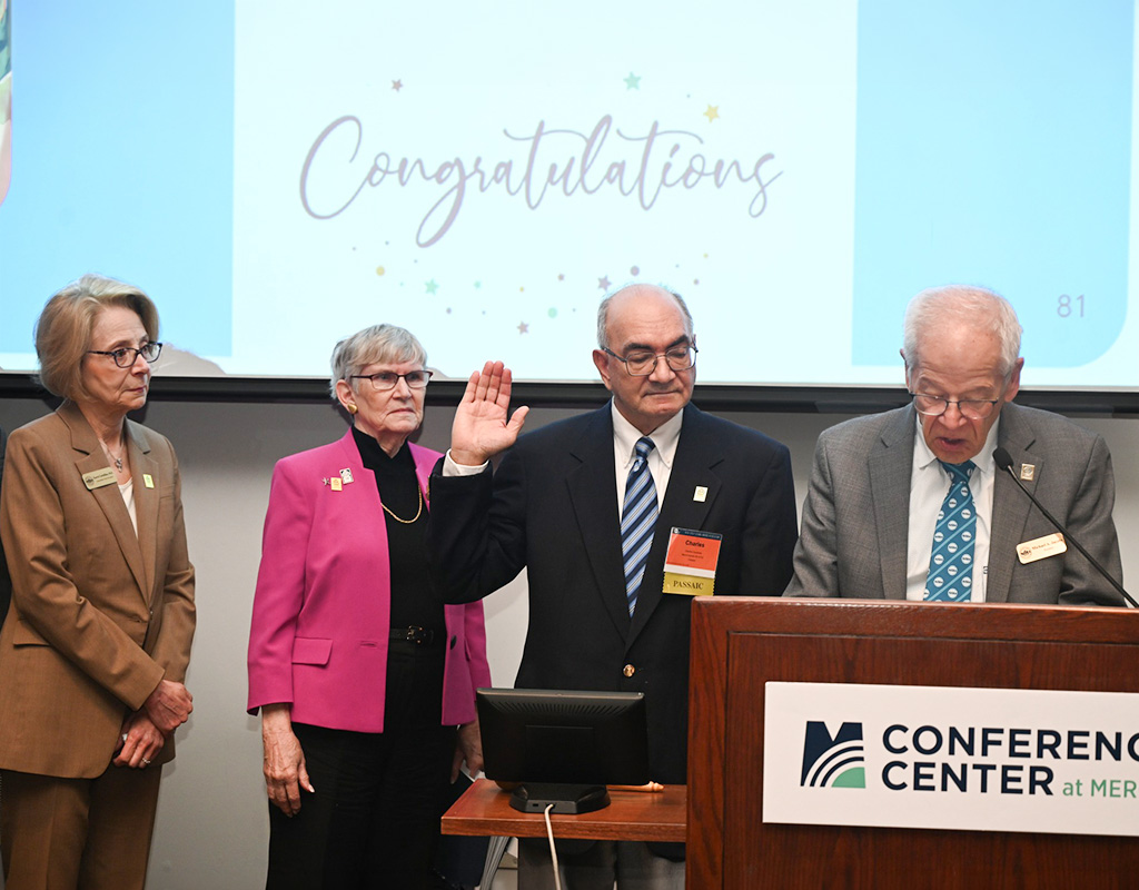 Michael Jacobs speaks while Charles Caravallo is sworn in, standing at a podium. Dr. Karen Cortellino and Irene LeFebvre stand beside. Congratulations is displayed on the screen behind.