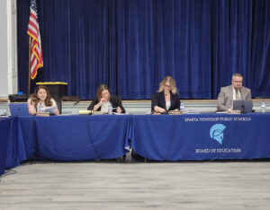 Sparta BOE members sit at a head table with microphones. The tablecloth is blue with the Sparta logo
