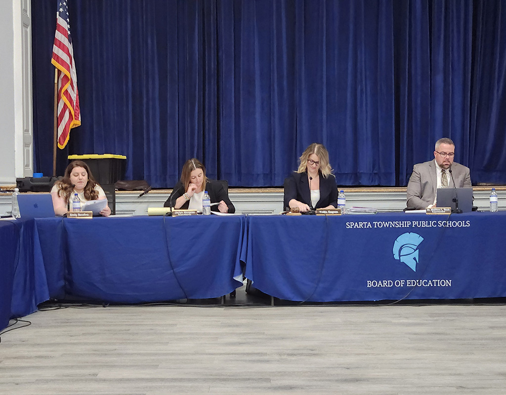 Sparta BOE members sit at a head table with microphones. The tablecloth is blue with the Sparta logo