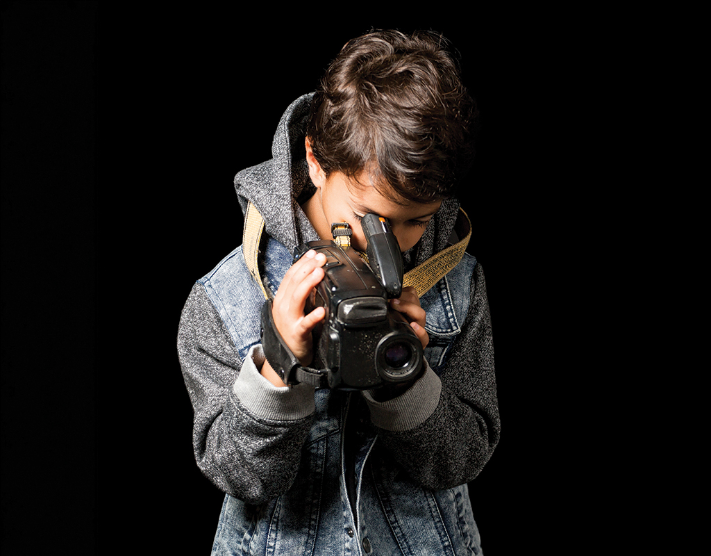 Middle school-aged student in a hoodie holding a video camera to his face pointing down in front of a black background