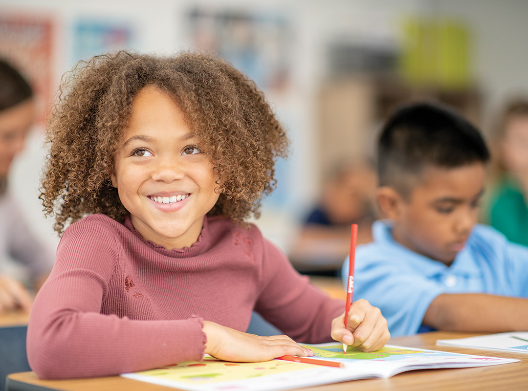 two grade school students sitting at a desk writing. one is smiling and the other is looking down intent