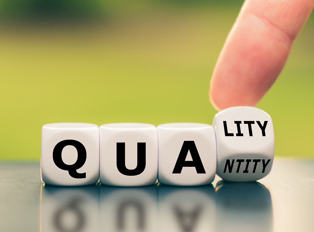 close up of letter blocks sitting on a table spelling out qua with the last block showing two sides one wiht lity the other ntity and a finger turning it. the background is a blurry green abstract